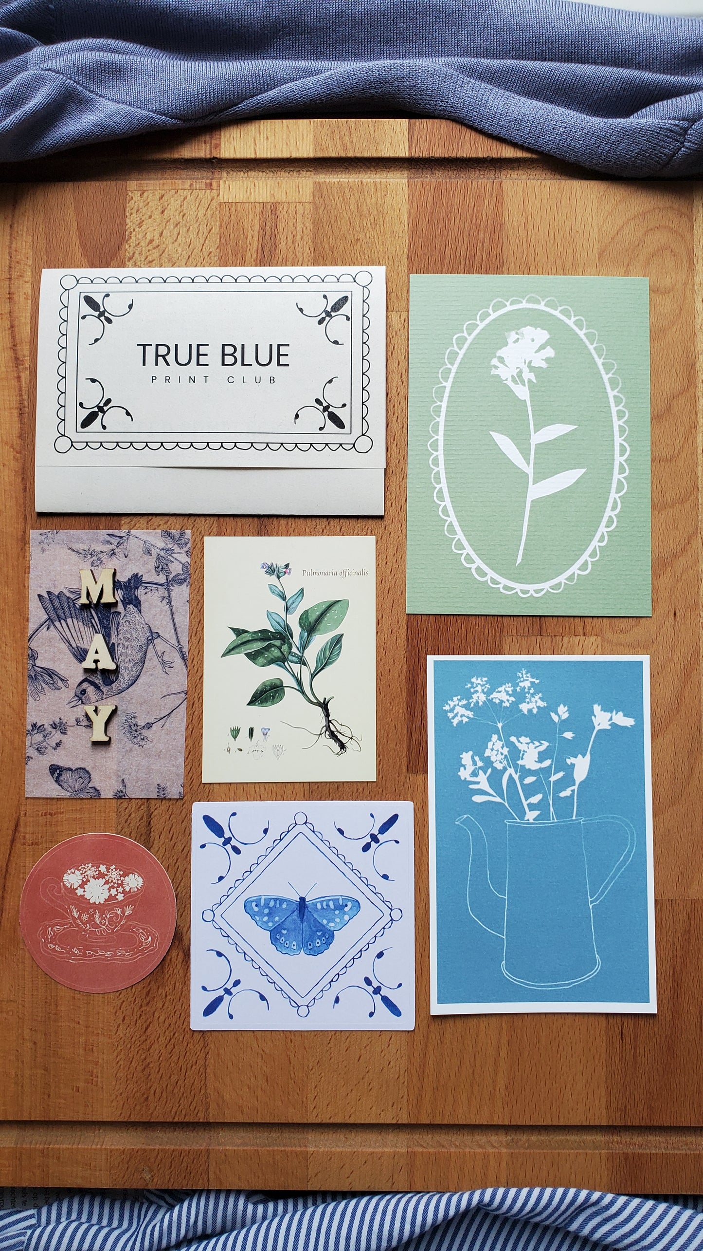 A curated flat-lay of six art prints and a small round card arranged on a wooden surface, flanked by blue striped fabric on both sides. The prints feature botanical and geometric designs in muted pastel tones including sage green, sky blue, cream, lavender, and dusty pink.