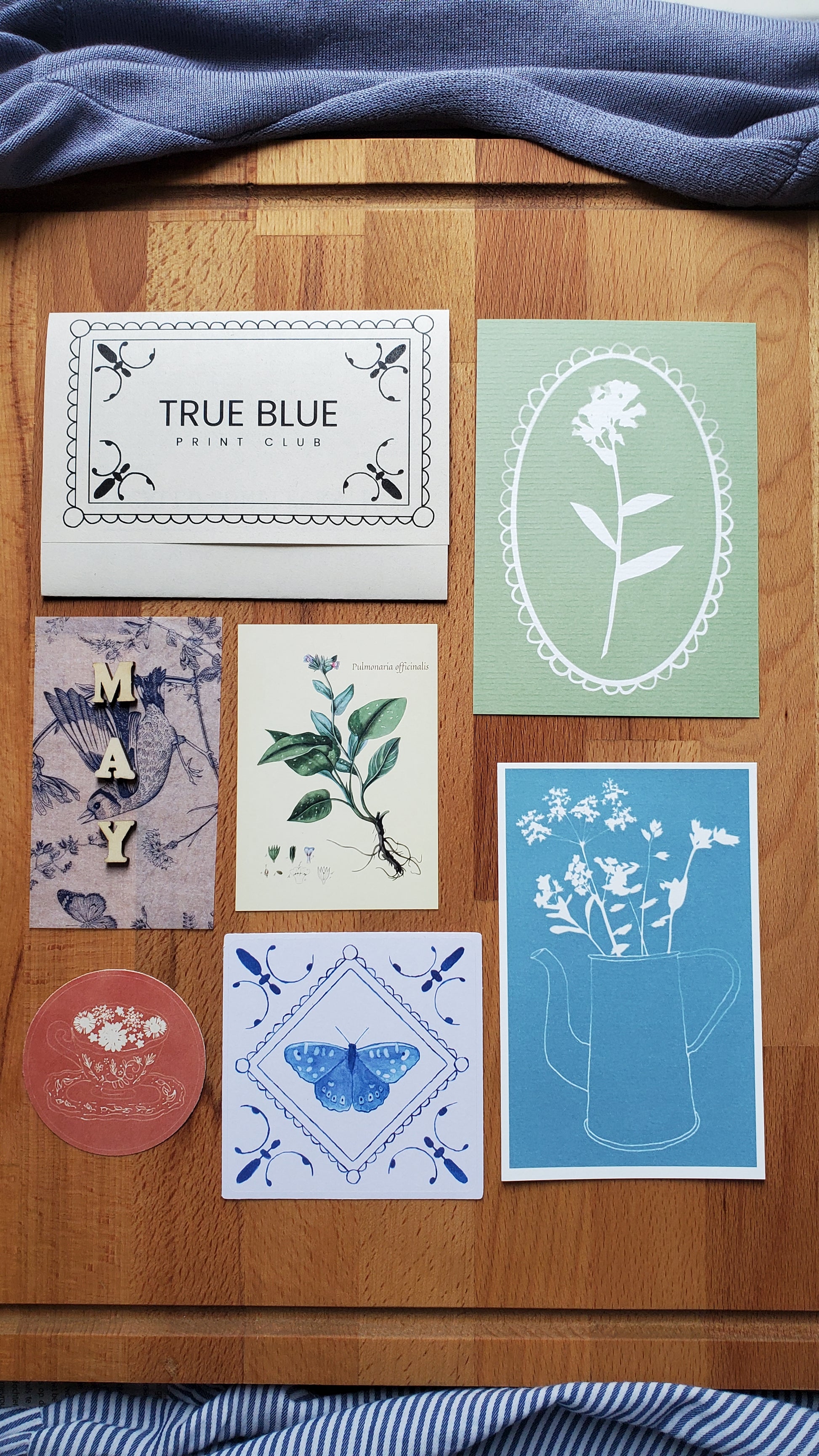 A curated flat-lay of six art prints and a small round card arranged on a wooden surface, flanked by blue striped fabric on both sides. The prints feature botanical and geometric designs in muted pastel tones including sage green, sky blue, cream, lavender, and dusty pink.